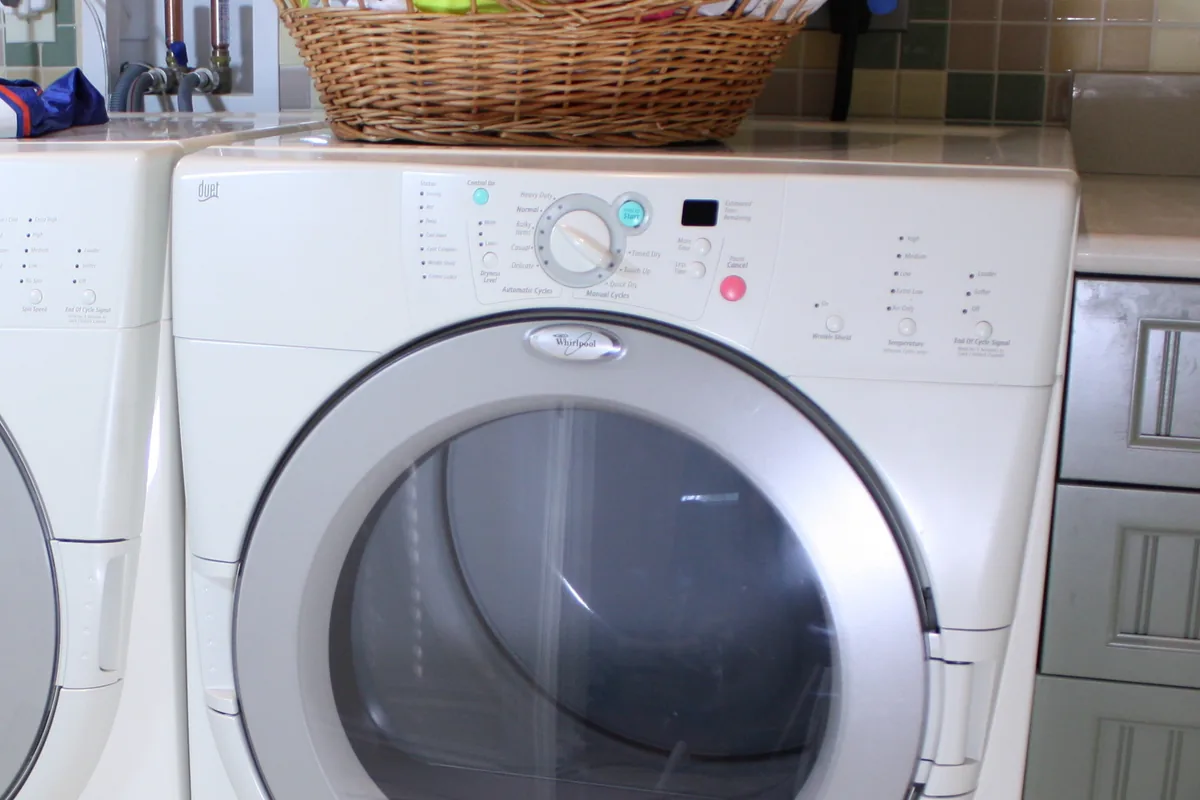 A white front-loading tumble dryer standing beside a washing machine in a domestic laundry room, with a wicker basket of clothes on top.