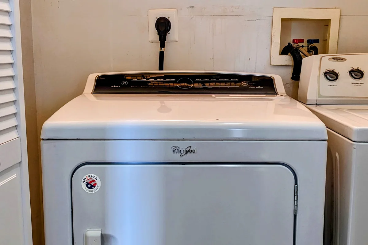 A white tumble dryer standing clear of a laundry room wall, with its power socket and the recessed service box for the neighbouring washing machine visible above it.