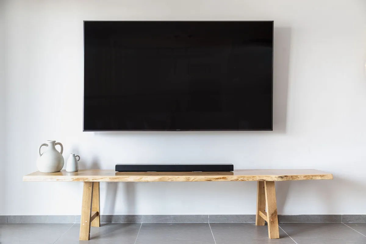 A large flat-screen television mounted on a plain white wall above a long, low wooden console holding a black soundbar and two ceramic jugs.