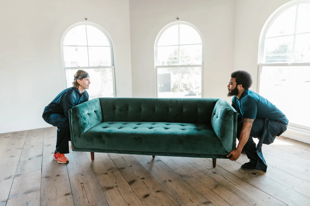 Two crew members crouched on either end of a sofa, lifting it together in an empty room.