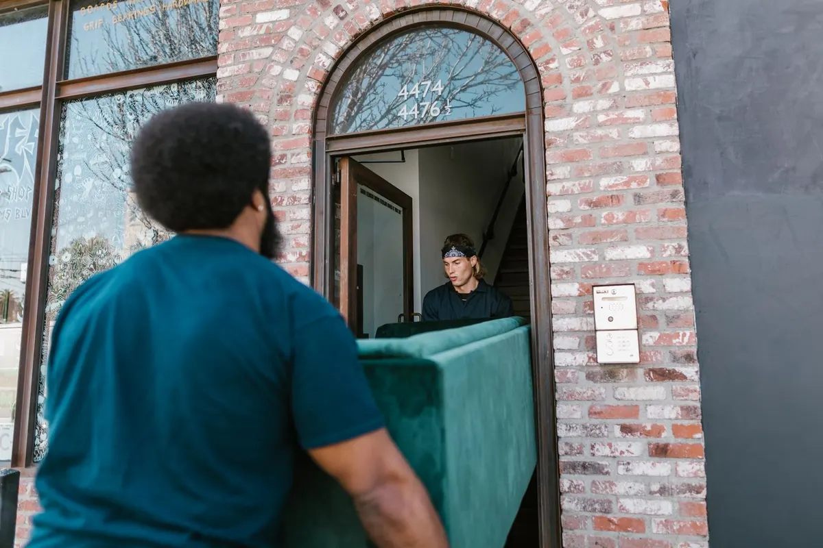 Two people carrying a green sofa on its side out through a narrow arched doorway, with a staircase visible in the hallway behind them.