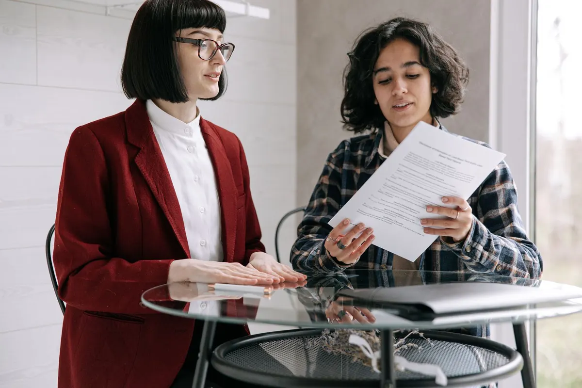 Two people at a glass table, one holding up a printed contract and reading it aloud while the other listens.