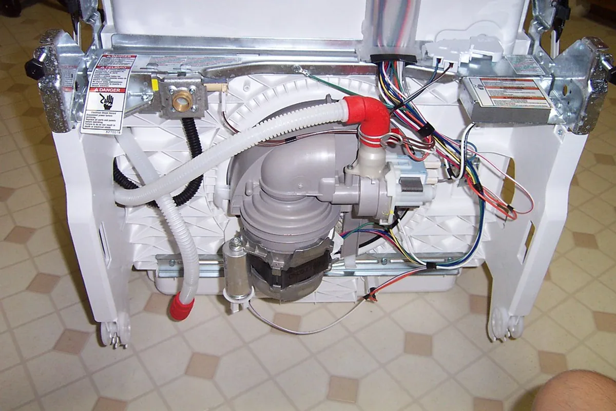 The exposed underside of a dishwasher lying on a tiled floor, showing the drain pump, corrugated hoses and coloured wiring beneath the base.