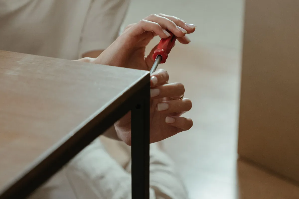 A person kneeling on the floor using a red-handled screwdriver on the fixing between a wooden table top and a black metal leg.
