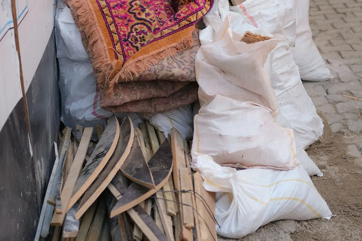 A pile of rolled-up old carpets, timber offcuts and tied woven rubble sacks stacked against a wall on a pavement, waiting to be collected.