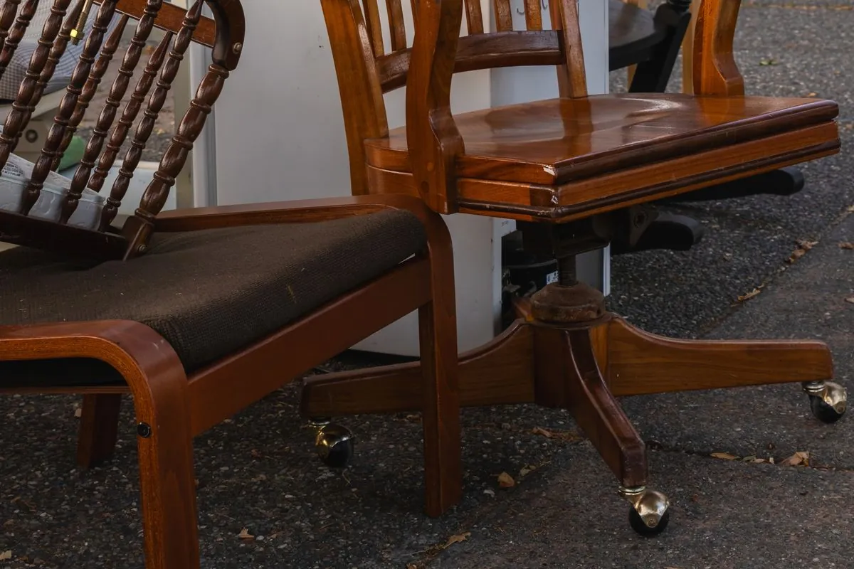 Used wooden chairs, including a swivel desk chair on castors and a bentwood chair with a worn fabric seat, standing on tarmac at an outdoor second-hand furniture stall.