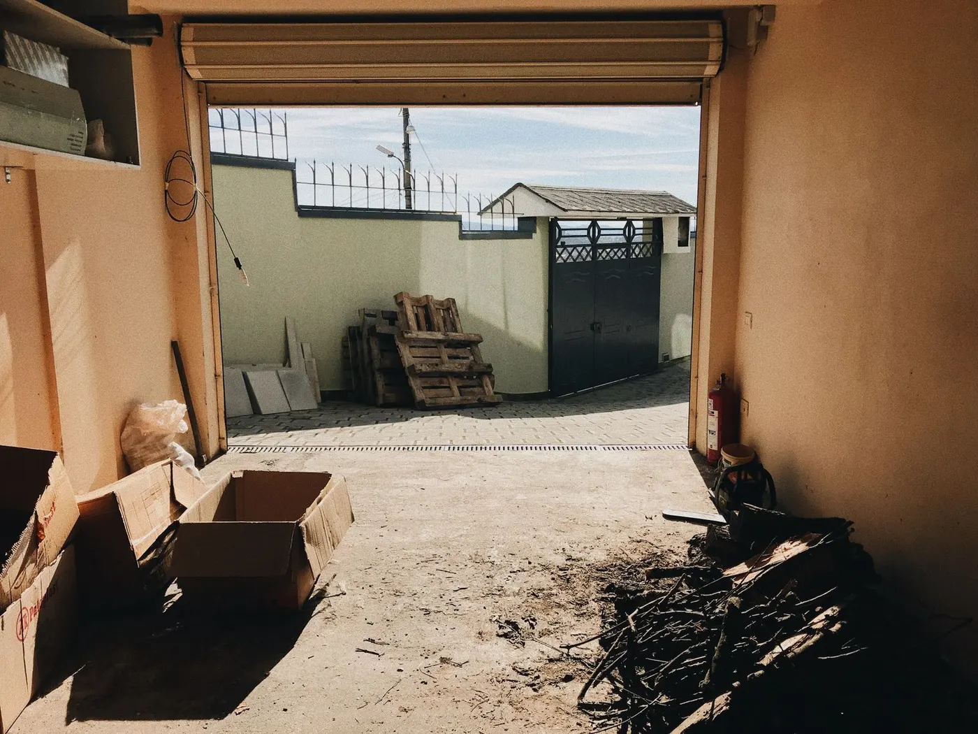 The interior of a villa garage looking out through an open roller door at a stack of wooden pallets and boxes in the courtyard.