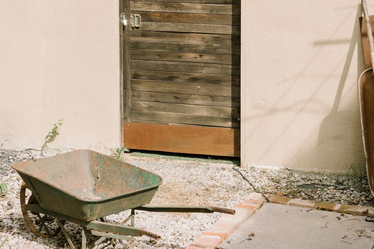 A weathered wooden door into an outdoor store in a cream rendered wall, with a rusted green wheelbarrow standing on gravel in strong sunlight.