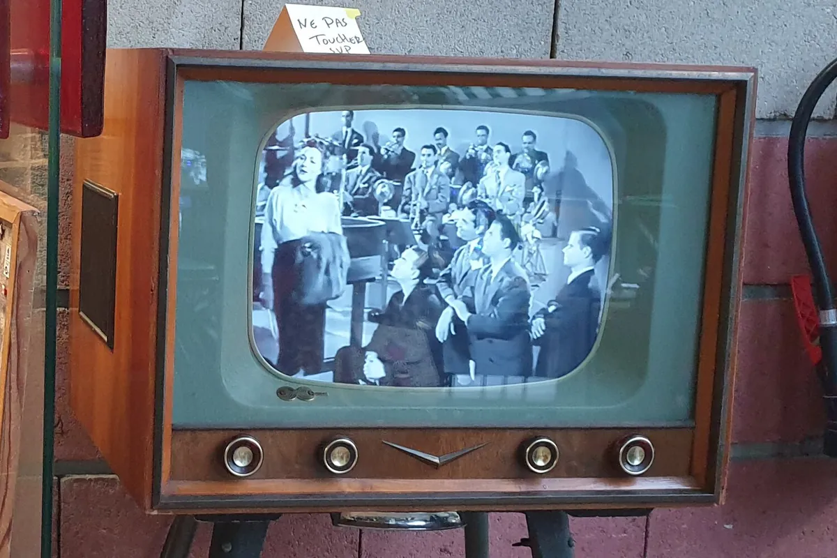 A 1950s wooden-cased television showing a black-and-white picture, displayed on a metal tripod stand against a brick wall.