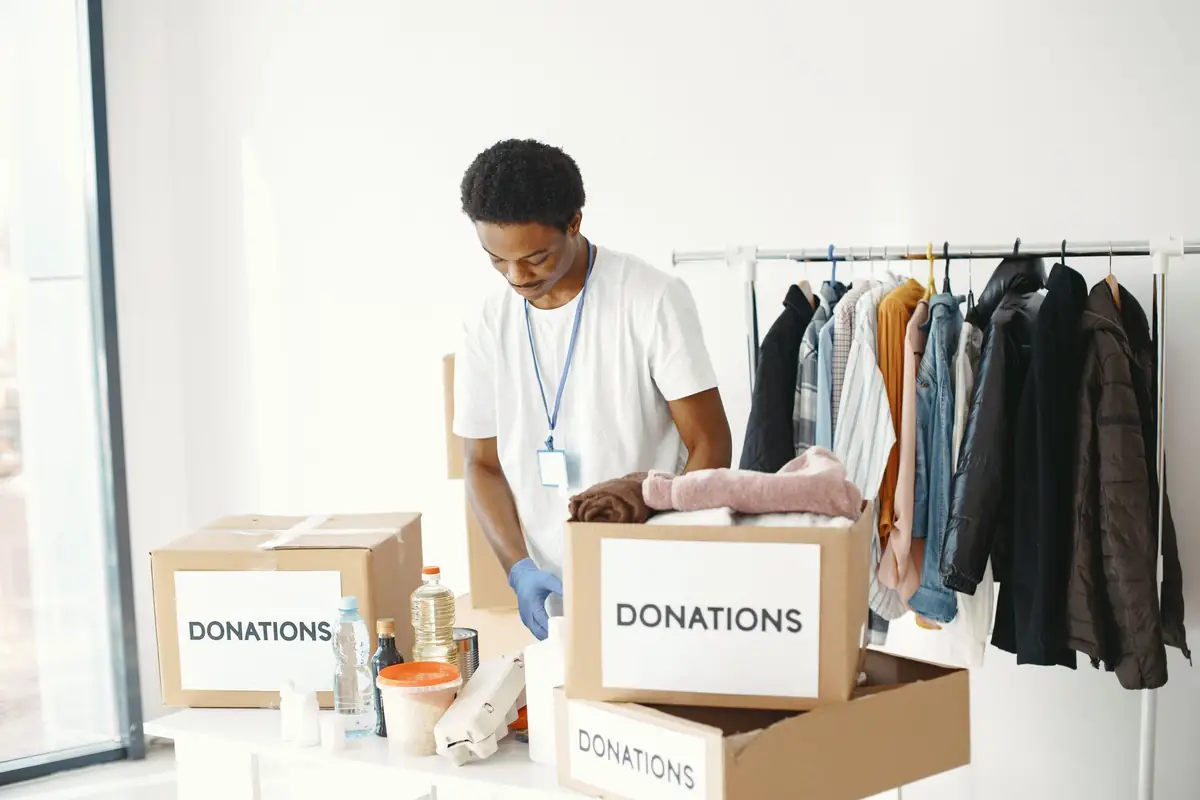A volunteer in gloves sorting goods into cardboard boxes marked Donations at a collection point, with a rail of donated coats behind him.