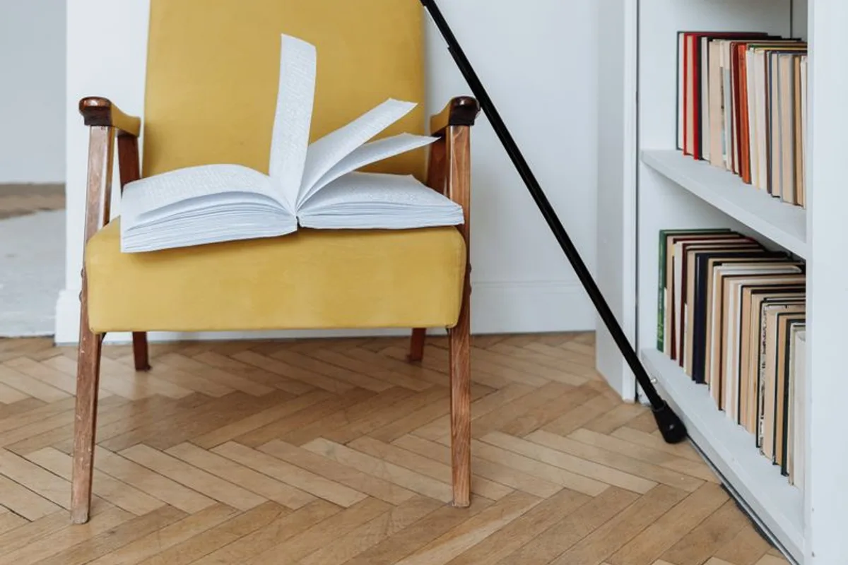 A wooden walking cane leaning against a yellow armchair beside a bookshelf full of books.
