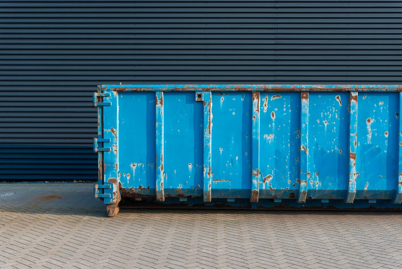 A large blue metal skip container standing on block paving against a dark corrugated-metal building wall.