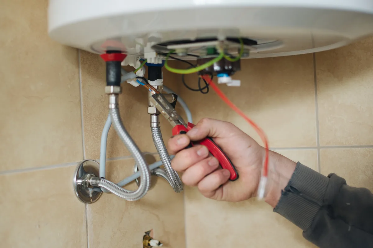 A technician using pliers on the flexible water hoses and wiring beneath a wall-mounted water heater.