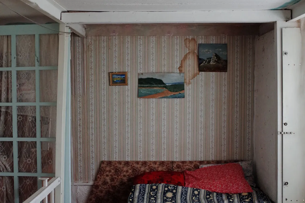 A brown water stain running down patterned wallpaper between two hanging pictures, directly above a bed pushed against the wall.
