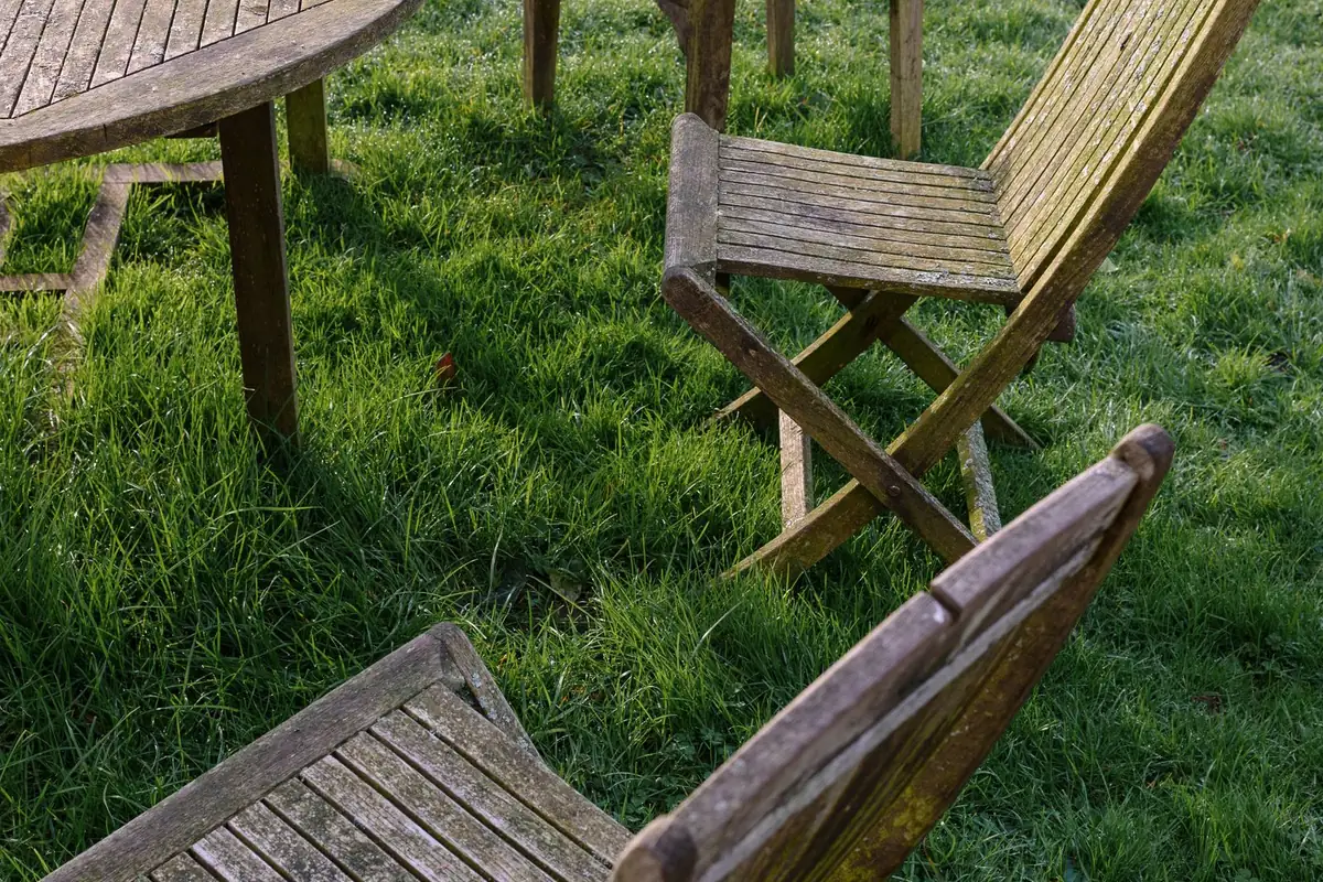 A round slatted timber garden table and folding chairs standing on grass, the wood weathered to a dry silver-grey with the grain raised.
