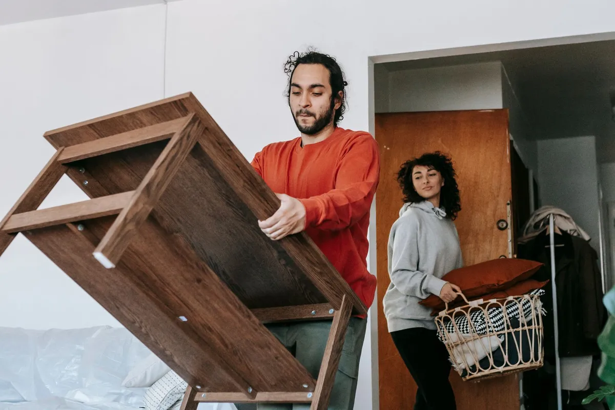 A man tilting a wooden shelf unit while a woman carries a basket of cushions towards the door of a bedroom with the bed covered in protective sheeting.