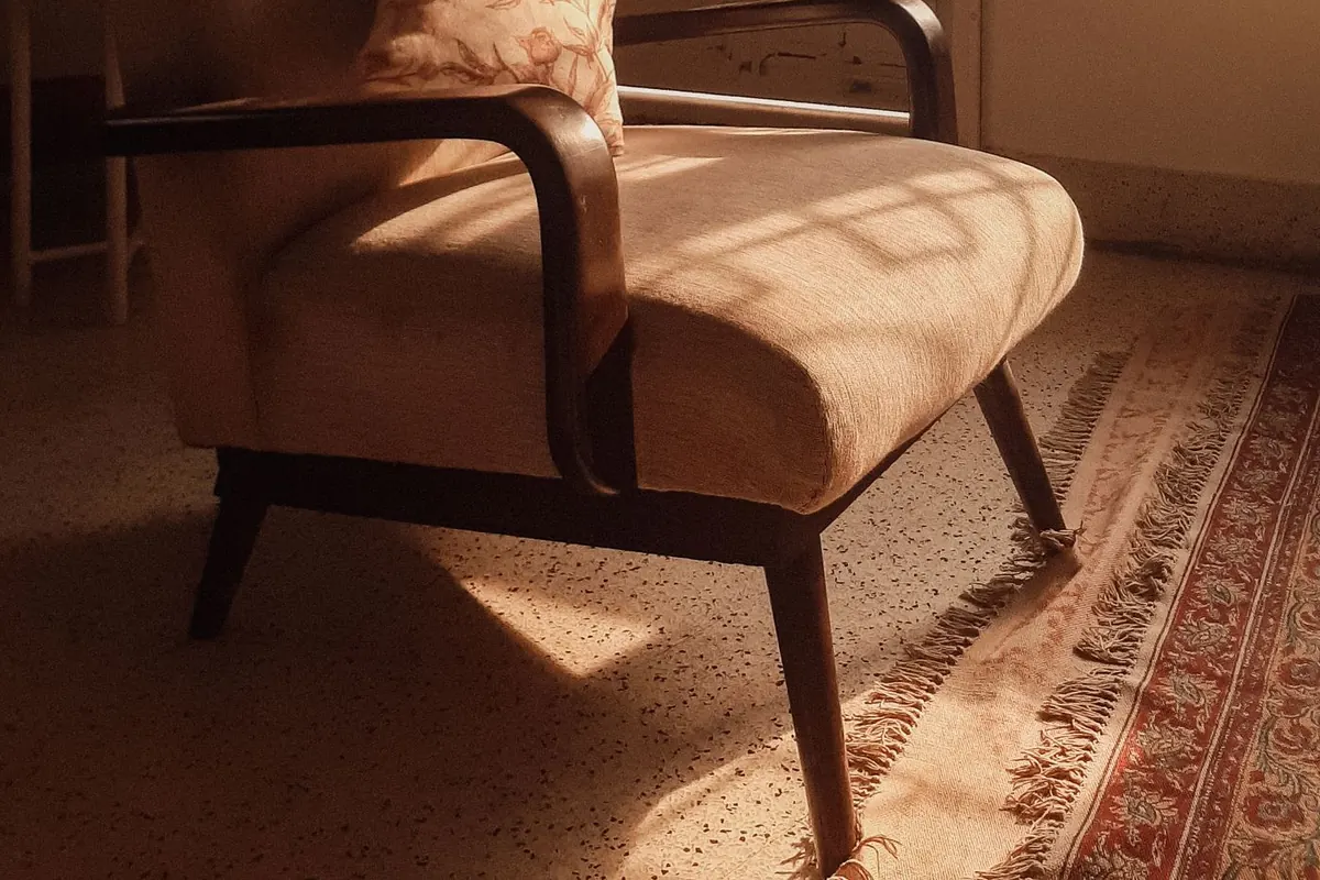 Low afternoon sunlight falling across the linen seat of an armchair with a curved dark wooden arm and splayed tapered legs, standing on the fringed edge of a patterned rug.