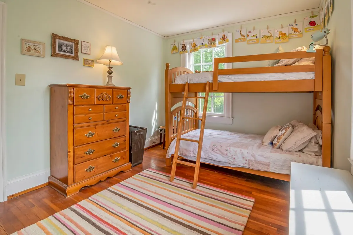 A pine bunk bed with a slatted guard rail and a leaning wooden ladder standing against the window wall of a child's bedroom, beside a tall chest of drawers.