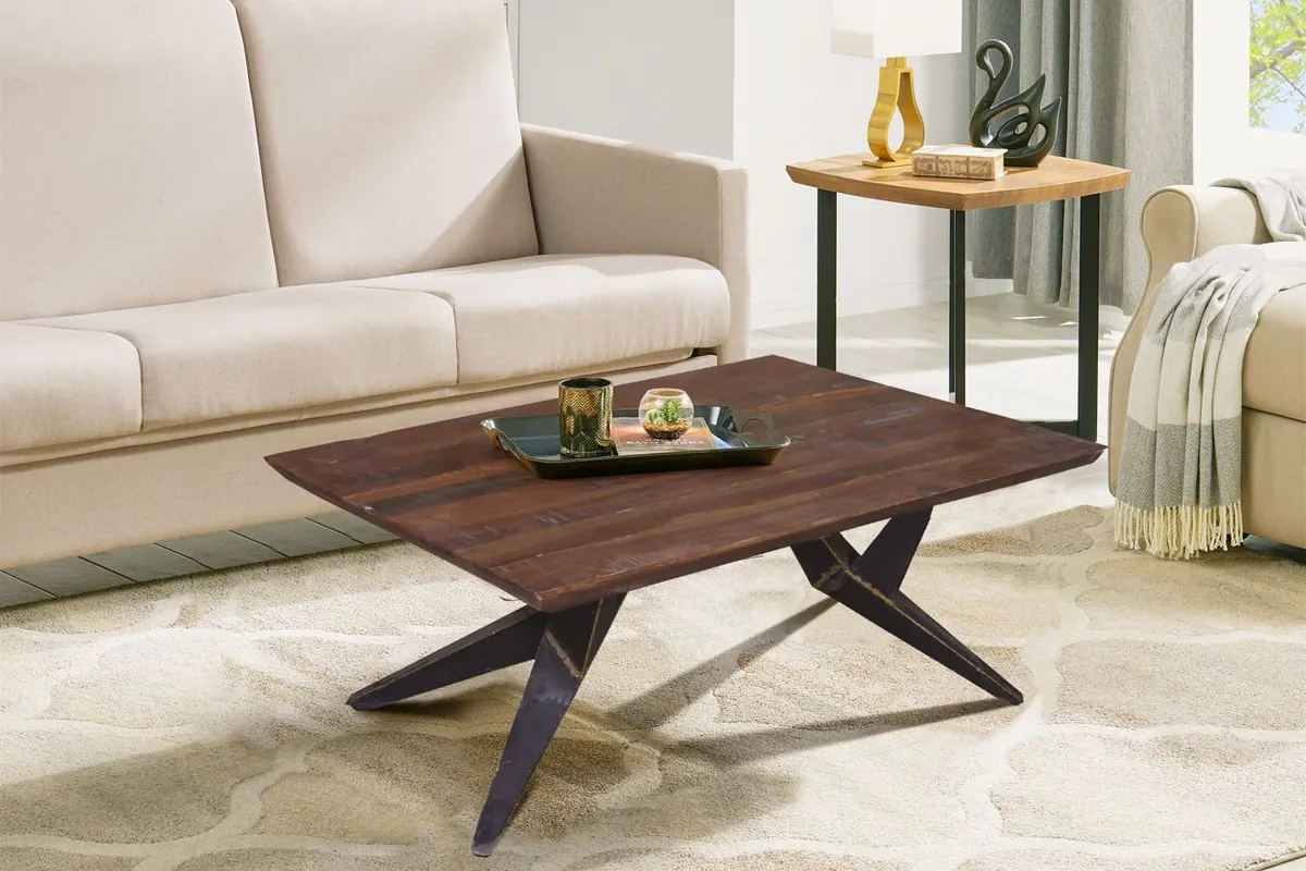 A rectangular dark wooden coffee table on splayed black metal legs standing on a patterned cream rug in front of a cream fabric sofa, with a side table and curtains behind.