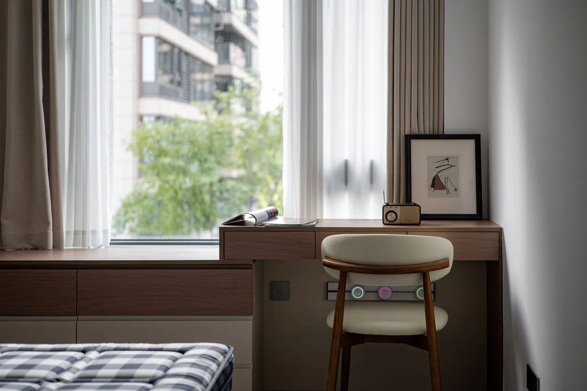 A wooden desk with drawers and a curved chair set against a bedroom window, with a neighbouring apartment tower and trees visible outside.