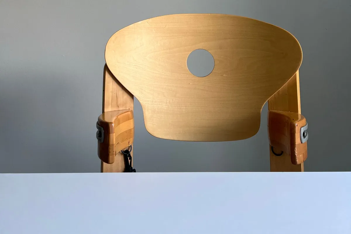 The curved plywood back of an adjustable wooden high chair seen from behind, with the harness strap fittings and height-adjustment bolts on each side.