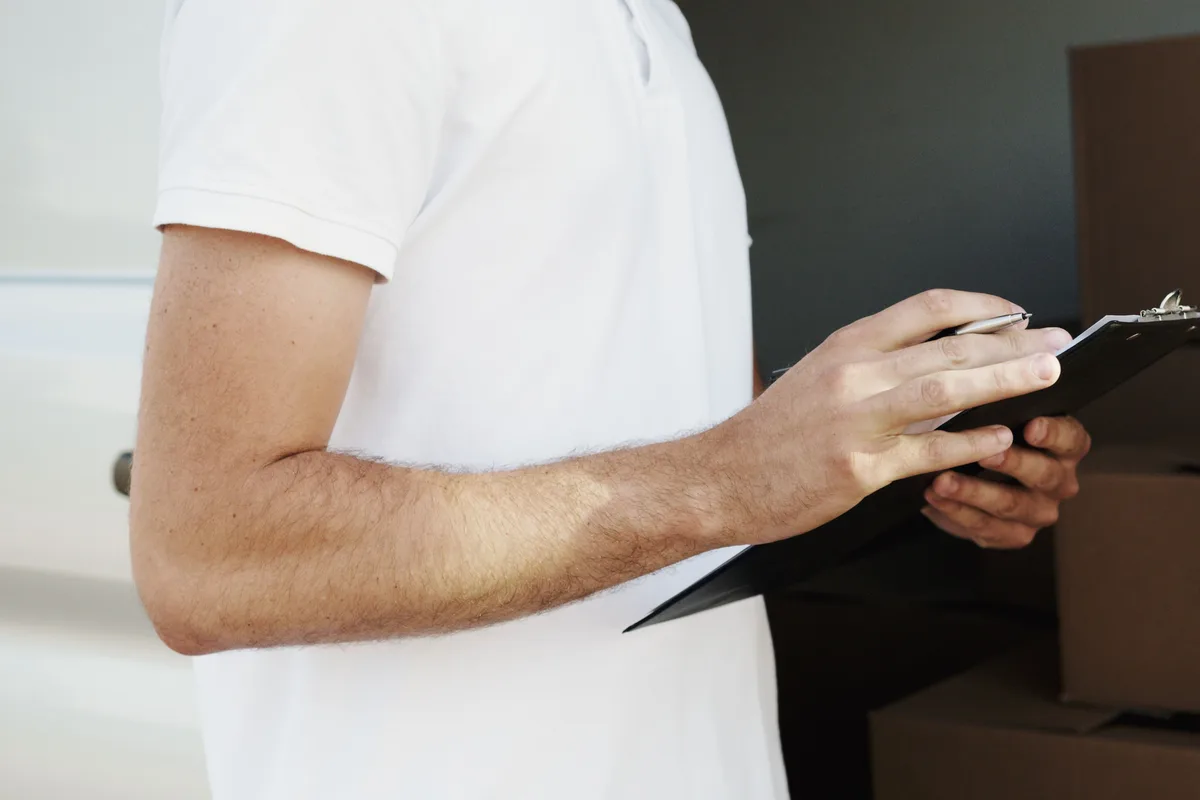 A worker in a white polo shirt writing on a clipboard beside a stack of cardboard boxes, with a van visible behind him.