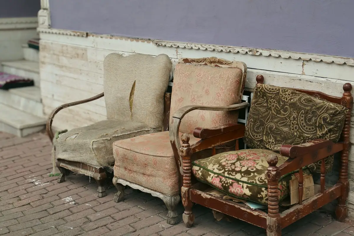 Three worn armchairs standing on a brick pavement against a wall, one with its seat collapsed and webbing hanging loose, another stripped back to its bare wooden frame.