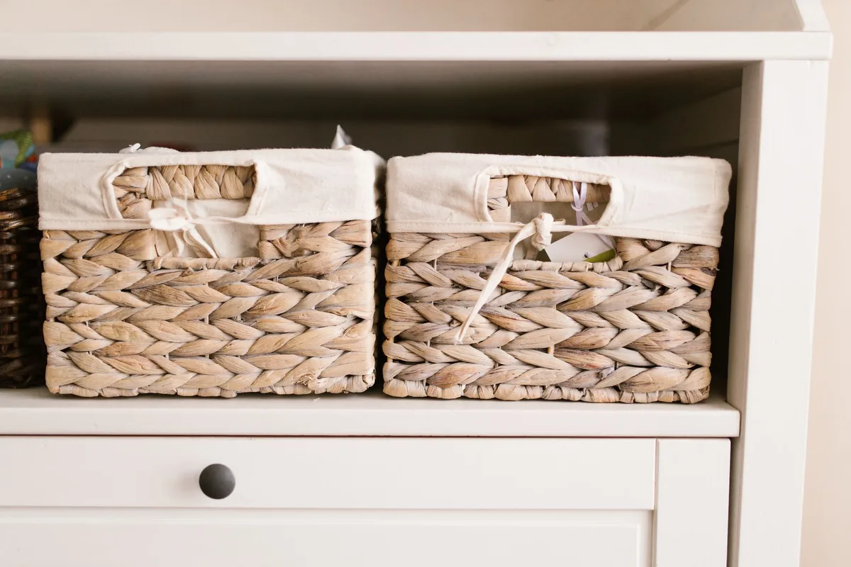 Two woven storage baskets with cream fabric linings sitting side by side on an open shelf above a white cabinet drawer.