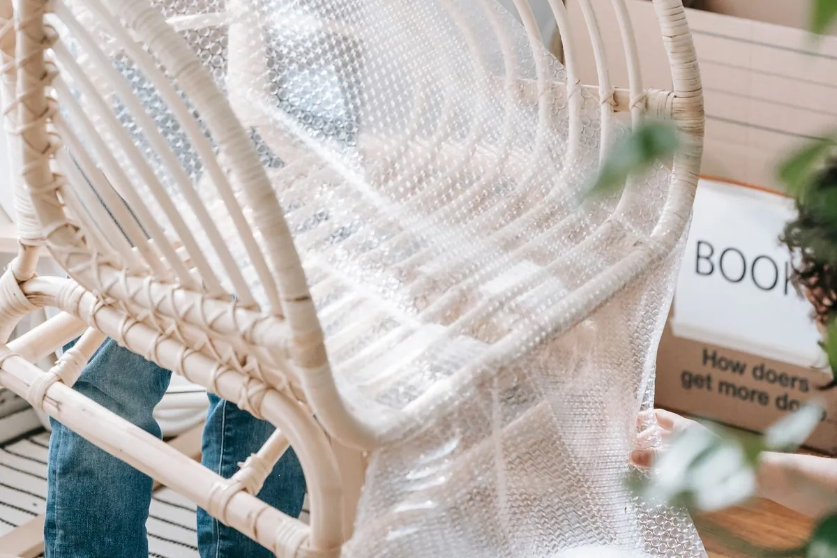 Bubble wrap being drawn over the back and seat of a rattan chair in a flat, with packed cartons stacked behind it.